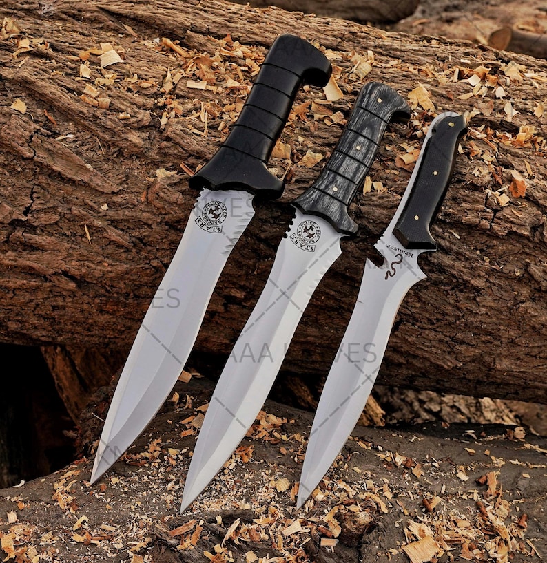 May include: Three daggers with silver blades and black handles are displayed on a wooden surface. The handles have different designs, including a curved shape and a textured grip. Each blade has a decorative emblem near the handle.