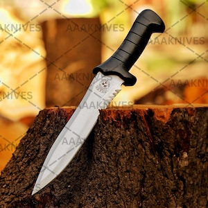 May include: A survival knife with a silver blade and a black handle. The knife has a serrated edge near the handle and a logo with stars. The knife is resting on a tree stump. The text "AAAKNIVES" is visible.