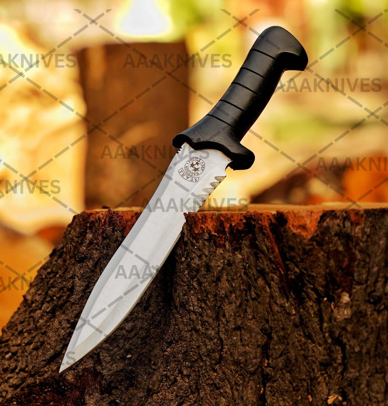 May include: A survival knife with a silver blade and a black handle. The knife has a serrated edge near the handle and a logo with stars. The knife is resting on a tree stump. The text "AAAKNIVES" is visible.
