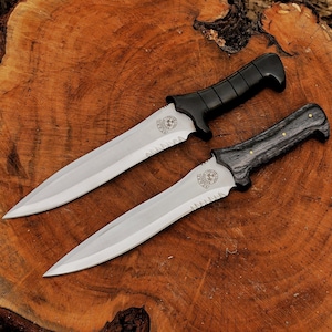 May include: Two tactical knives with serrated blades and black handles are displayed on a wooden surface. The blades are silver, and the handles have a textured design. One handle is black, and the other is gray with brass accents. The knives have a decorative emblem near the handle.
