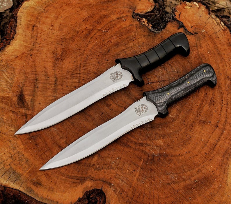 May include: Two tactical knives with serrated blades and black handles are displayed on a wooden surface. The blades are silver, and the handles have a textured design. One handle is black, and the other is gray with brass accents. The knives have a decorative emblem near the handle.