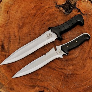 May include: Two knives with silver blades and black handles rest on a wooden surface. The larger knife has a serrated edge and a black, textured handle. The smaller knife has a smooth blade and a black handle with gold-colored rivets.