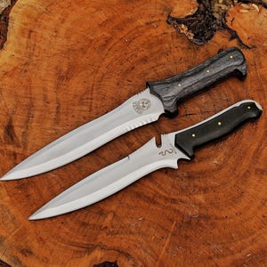 May include: Two knives with silver blades and dark wooden handles are displayed on a wooden surface. The larger knife has a textured handle and a serrated edge near the hilt. The smaller knife has a smooth, black handle.