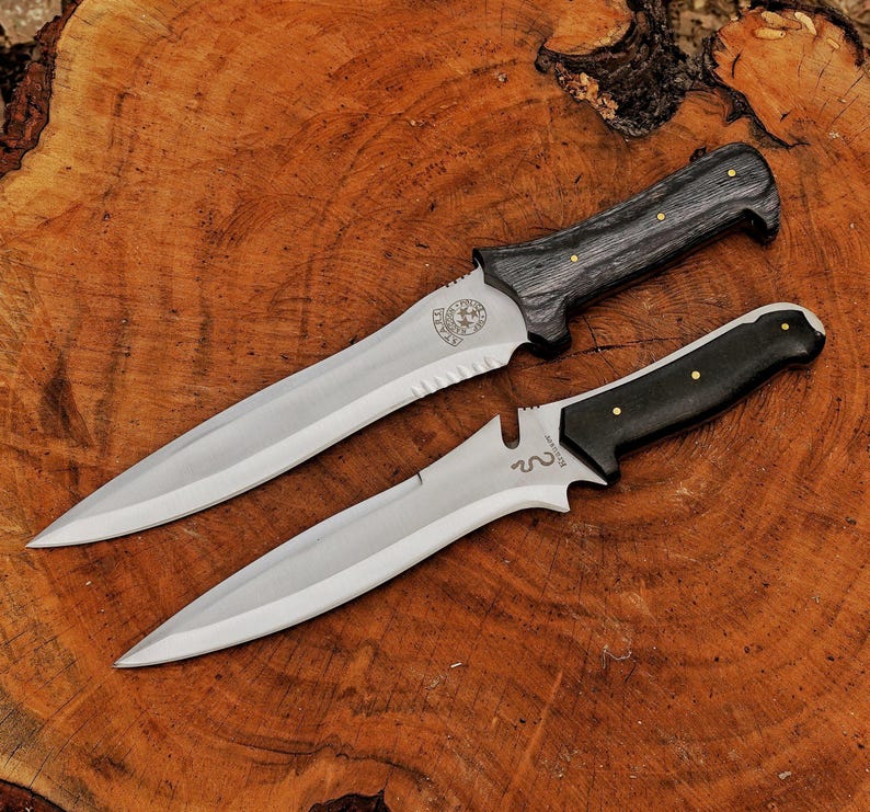 May include: Two knives with silver blades and dark wooden handles are displayed on a wooden surface. The larger knife has a textured handle and a serrated edge near the hilt. The smaller knife has a smooth, black handle.