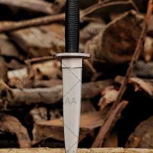 May include: A silver dagger with a black, ridged handle and a silver pommel. The blade is double-edged and tapers to a point. The dagger is displayed on a wooden surface against a backdrop of stacked logs. The blade has the letters "AA" etched on it.