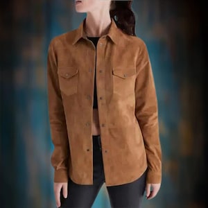 May include: A brown suede shirt with two chest pockets and button closures. The long-sleeved shirt has a collared neckline and a slightly curved hemline. The shirt is worn over a black top and paired with black pants.