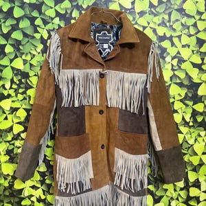 Könnte beinhalten: Braune Wildlederjacke mit Fransen. Die Jacke hat ein Patchwork-Design mit verschiedenen Braun- und Beigetönen. Sie hat einen Kragen, einen Knopfverschluss und Taschen. Die Fransen sind hellbeige.