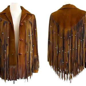 May include: A brown suede jacket with long fringe detailing and pearl-like embellishments. The jacket features a collared neckline, three buttons, and fringe along the sleeves, front, and back. The jacket is displayed on a mannequin.
