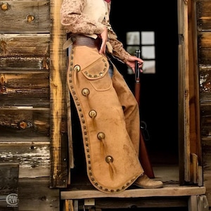 May include: Tan leather chaps with decorative brass studs and accents. The chaps feature a pocket and fringe details. The image shows a rustic, western-themed aesthetic, with a wooden door and a rifle in the background.