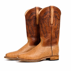 Könnte beinhalten: Ein Paar hell- und dunkelbraune Cowboystiefel mit einem strukturierten, genarbten Lederfuß und einem glatten Lederschaft. Der obere Schaft ist mit aufwendigen dunkelbraunen Nähten verziert und verfügt über zwei vertikale Anziehschlaufen.