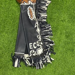 Peut inclure: Chaps en cuir noir avec des accents décoratifs blancs et marron et des franges. Les chaps ont une ceinture et un motif décoratif avec les lettres "ECS" en blanc. Les chaps sont sur une surface verte.