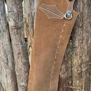 May include: Brown leather chaps with decorative stitching and silver hardware. The chaps are hanging on a wooden fence. The top portion features a lighter brown leather with a decorative design and a buckle strap.