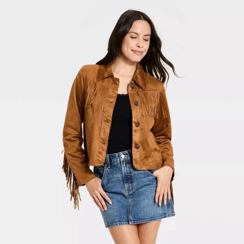 May include: A brown suede jacket with fringe detailing across the chest, shoulders, and sleeves. The jacket has a button-up front and a collared neckline. The model is wearing a black top and a denim skirt.