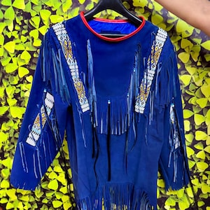 Puede incluir: Una camisa de ante azul con mangas largas, flecos y detalles de cuentas. La camisa tiene un ribete rojo alrededor del cuello y está colgada de una percha negra. Las mangas y la parte delantera están adornadas con largos flecos.