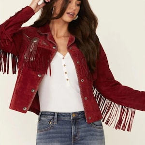 May include: A burgundy suede jacket with long fringe detailing on the sleeves and chest. The jacket features decorative embellishments and button closures. The model is wearing a white top and blue jeans.