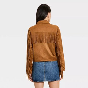 May include: A brown suede jacket with fringe detailing across the back and sleeves. The jacket has a collared neckline and long sleeves. The model is wearing a denim skirt. The jacket is a western-inspired style.