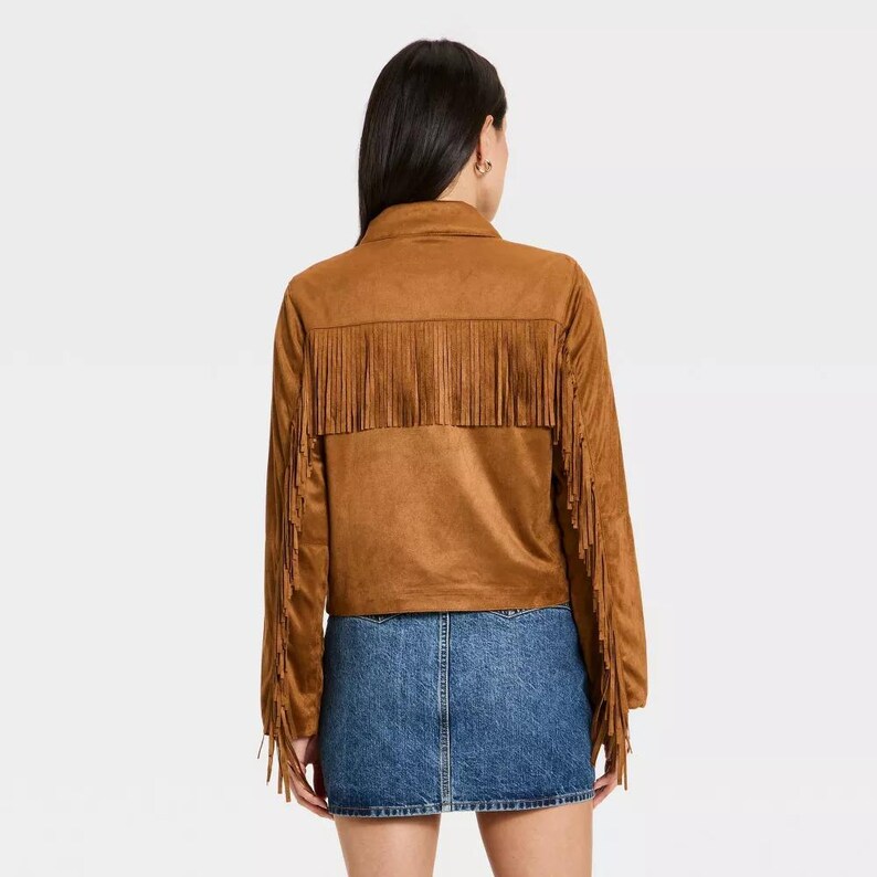 May include: A brown suede jacket with fringe detailing across the back and sleeves. The jacket has a collared neckline and long sleeves. The model is wearing a denim skirt. The jacket is a western-inspired style.