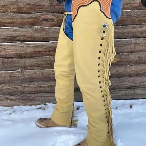 May include: Tan leather chaps with fringe detail and a decorative top. The chaps are worn over blue jeans and brown boots. A silver ring is attached to the chaps. The background is a wooden wall and snow.