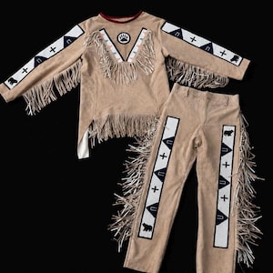 May include: A tan long-sleeve shirt and matching pants set featuring long fringe trim. The shirt has a V-neck with a bear paw print, while both pieces display white panels with blue geometric shapes, black bear silhouettes, and cross symbols.