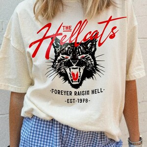 May include: Cream-colored t-shirt featuring a black cat graphic with red eyes and the text "The Hellcats" in red script. Below the cat, the shirt reads "Forever Raisin Hell - Est. 1978". The shirt is paired with blue and white checkered shorts.