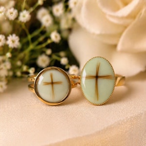 May include: Two gold rings with light green oval and round settings. Each setting features a cross design made of brown hair. The rings are displayed on a cream-colored surface, with white flowers in the background.