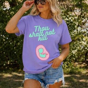 以下が含まれることがあります： ミントグリーンの「Thou shalt not kill」という文字が入ったライラック色のTシャツ。ピンクとグリーンの胎児のグラフィックが特徴です。デニムのショートパンツとサングラスを着用しています。