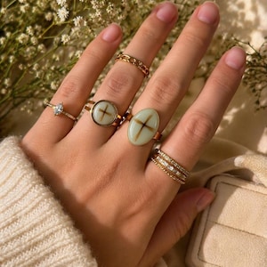 May include: A hand wearing several gold rings. Rings include a chain link style, a ring with a cross design, a ring with an oval-shaped stone with a cross design, a ring with a diamond-shaped stone, and a ring with multiple bands of small stones.