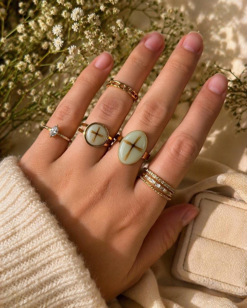 May include: A hand wearing several gold rings. Rings include a chain link style, a ring with a cross design, a ring with an oval-shaped stone with a cross design, a ring with a diamond-shaped stone, and a ring with multiple bands of small stones.