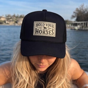 May include: Black trucker cap with a mesh back and a rectangular patch. The patch displays the text "HOLD YOUR HORSES" and a horse illustration. The cap is worn by a person with blonde hair.