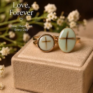 May include: Two gold rings with light green oval and round settings, each featuring a cross design made of brown hair. The rings sit on a beige velvet box, with the words "Love, Forever" and "Wear Their Love Every Day" in the background.