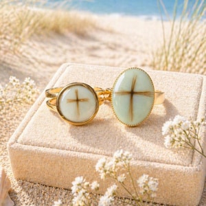 May include: Two gold rings with cross designs. One ring has a round face, the other an oval. The crosses are made of brown material set against a white and light green background. The rings are displayed on a beige surface with a beach background.