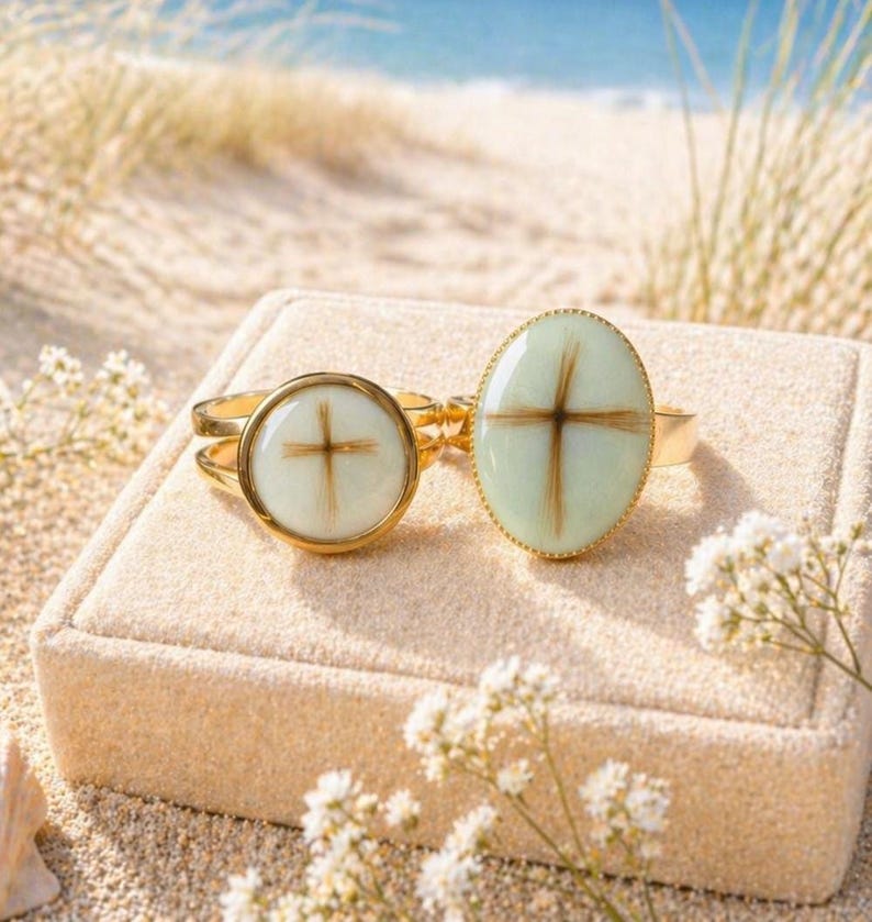 May include: Two gold rings with cross designs. One ring has a round face, the other an oval. The crosses are made of brown material set against a white and light green background. The rings are displayed on a beige surface with a beach background.