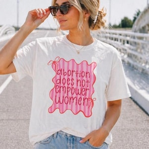 Puede incluir: Camiseta de color crema con un diseño de rayas rosas y blancas que dice "Abortion does not empower women." La camiseta tiene mangas cortas y un corte holgado. El texto está en negrita, con una fuente sans-serif.