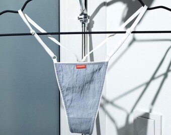 May include: A gray thong-style undergarment with white straps, hanging from a black hanger. The garment has a rectangular red tag with white text. The background is a white wall with a light fixture.