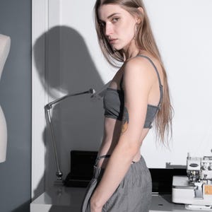 May include: A grey bra top and matching grey trousers. The model is standing in front of a white wall with a sewing machine and a desk. The model has long hair and is wearing hoop earrings.