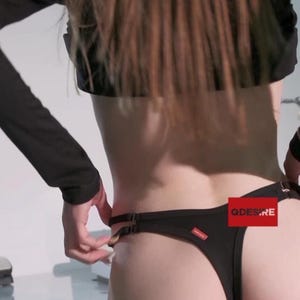 Könnte beinhalten: Schwarzer String-Tanga mit dünnem Bund und kleinem rotem Etikett. Der Text "QDESIRE" ist auf einem roten rechteckigen Etikett zu sehen. Das Kleidungsstück wird von einer Person angepasst.
