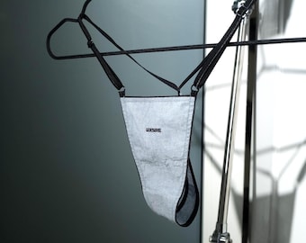 May include: A light gray thong with black trim and straps, hanging from a black hanger. The word "DESIRE" is embroidered on the front. The garment is made of a soft, textured fabric.