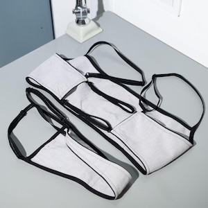 May include: Several pairs of light gray underwear with black trim and straps are arranged on a gray surface. The underwear has a simple, minimalist design. The straps are adjustable.