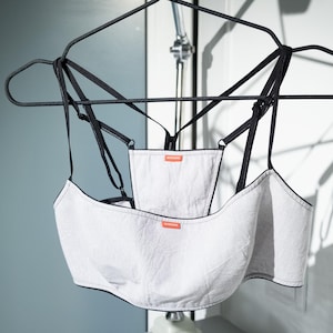 May include: A white and black lingerie set hanging on a black hanger. The set features a bralette and a high-waisted bottom, both with black trim and straps. Small orange tags are visible.