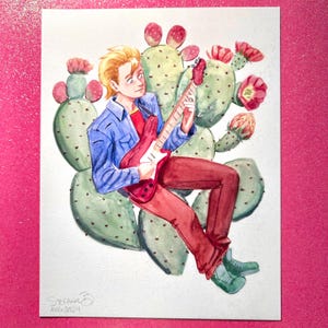 May include: Watercolor illustration of a person playing a red electric guitar, seated among green cacti with pink flowers. The person is wearing a blue jacket, red shirt, and red pants. Signed "Stefanie" and dated "AUG 2024".