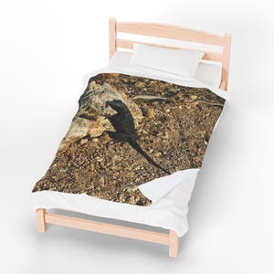 May include: A black lizard on a rocky surface is featured on a duvet cover. The lizard is in sharp focus against a backdrop of brown and tan rocks and pebbles. The duvet cover is on a wooden bed frame with a white pillow.