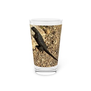 May include: A clear glass featuring a detailed image of a black lizard on a rocky surface. The lizard is positioned diagonally, with its tail extending downwards. The background shows a mix of brown and tan tones.