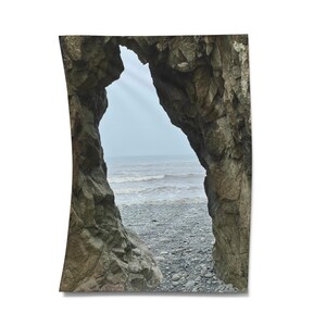 Peut inclure: Photographie d'une arche rocheuse naturelle encadrant une vue sur l'océan. L'arche est composée de roches brutes, grises et brunes, avec la mer et une plage de galets visibles à travers l'ouverture. Le ciel est nuageux.