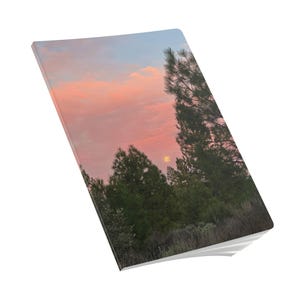 May include: A notebook with a landscape image of a sunset. The image shows a pink and blue sky, green trees, and the sun. The notebook's pages are visible on the bottom edge.