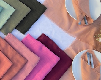 May include: A collection of colorful cloth napkins in various shades, including blue, green, orange, pink, and burgundy, arranged on a white tablecloth. Two plates with folded napkins and silverware are set on a table.