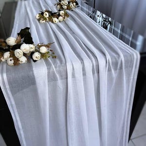 Może przedstawiać: Biały, przezroczysty bieżnik na stół z dwoma białymi kompozycjami kwiatowymi na ciemnym drewnianym stole.