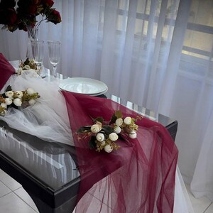 May include: A burgundy and white tulle table runner with white flowers and greenery. The runner is on a black table with a white plate and two wine glasses.