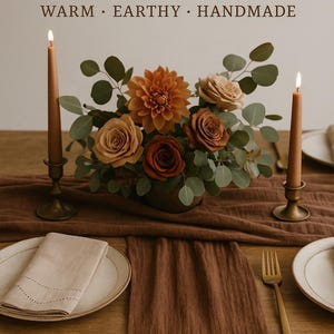 Pode incluir: Uma mesa posta com um caminho de mesa de gaze cor de mocha, um centro de mesa floral com rosas laranja e castanhas, e velas acesas em castiçais de latão. O texto "MOCHA MOUSSE GAUZE TABLE RUNNER" está no topo.