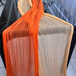 Op de afbeelding: Een geplooide tafelloper in oranje en beige tinten. De transparante stof drapeert over een donker oppervlak en creëert een vloeiend, gestructureerd effect. De kleuren van de loper gaan over van een levendig oranje naar een zacht beige.
