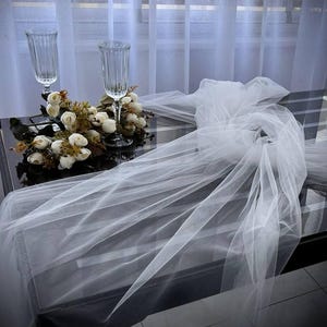 Puede incluir: Un camino de mesa de tul blanco con una guirnalda floral y dos copas de vino de vidrio transparente sobre una mesa negra.